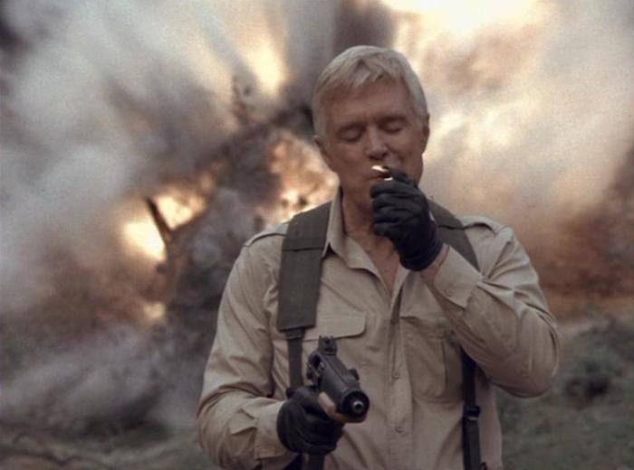 Hannibal from A-Team lighting a cigar and holding a gun while a vehicle explodes in the background
